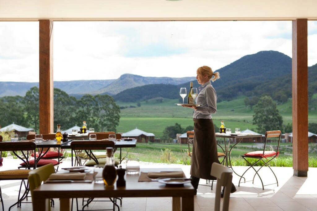 A server carrying a bottle of wine with two glasses in the alfresco dining area of Wolgan Valley Resort.