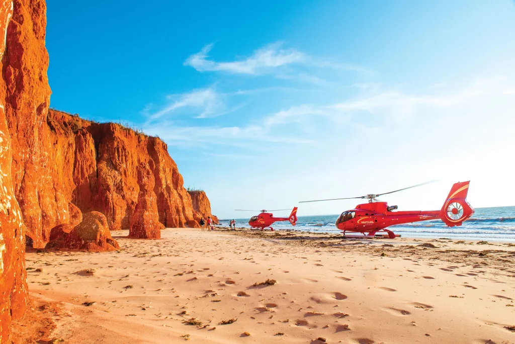 Two red helicopters landed on a remote beach in the Kimberley region of Western Australia, landing alongside the ocean and red pindan cliff face