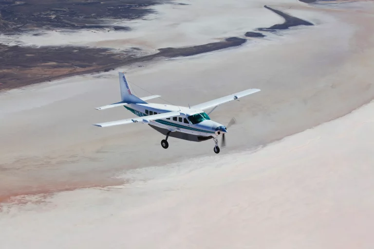 A Cessna Caravan aircraft with propeller in motion flying across a salt lake