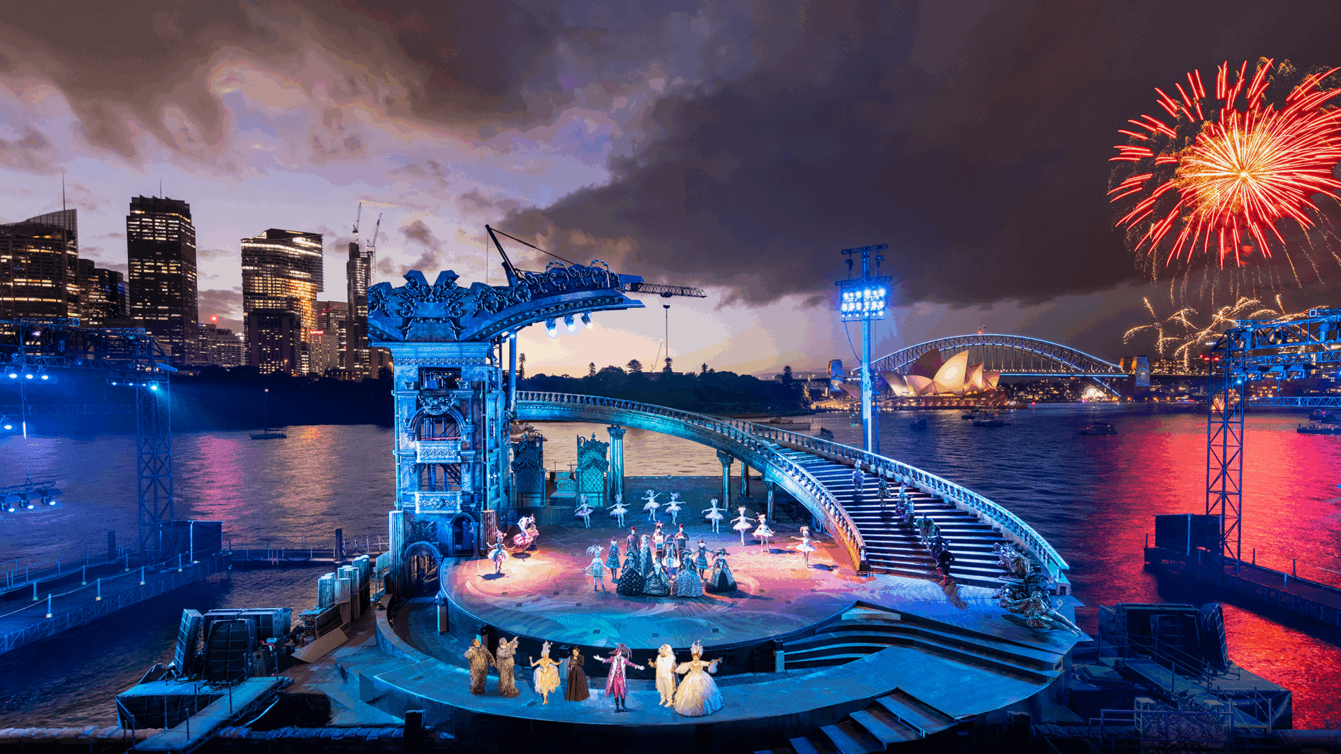 An opera stage on the side of a body of water with a bridge, an opera house and fireworks in the background.
