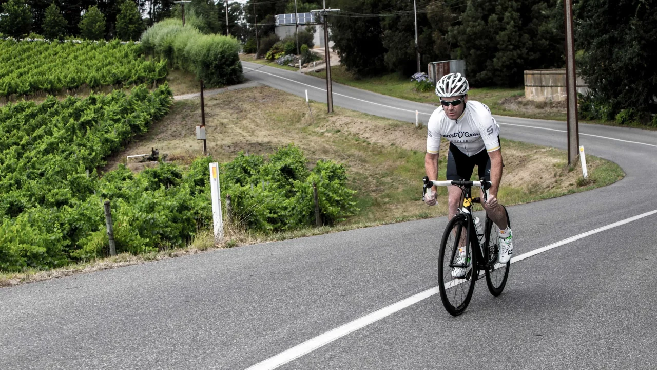 Stuart O’Grady cycling on a winding road in the Adelaide Hills alongside grapevines